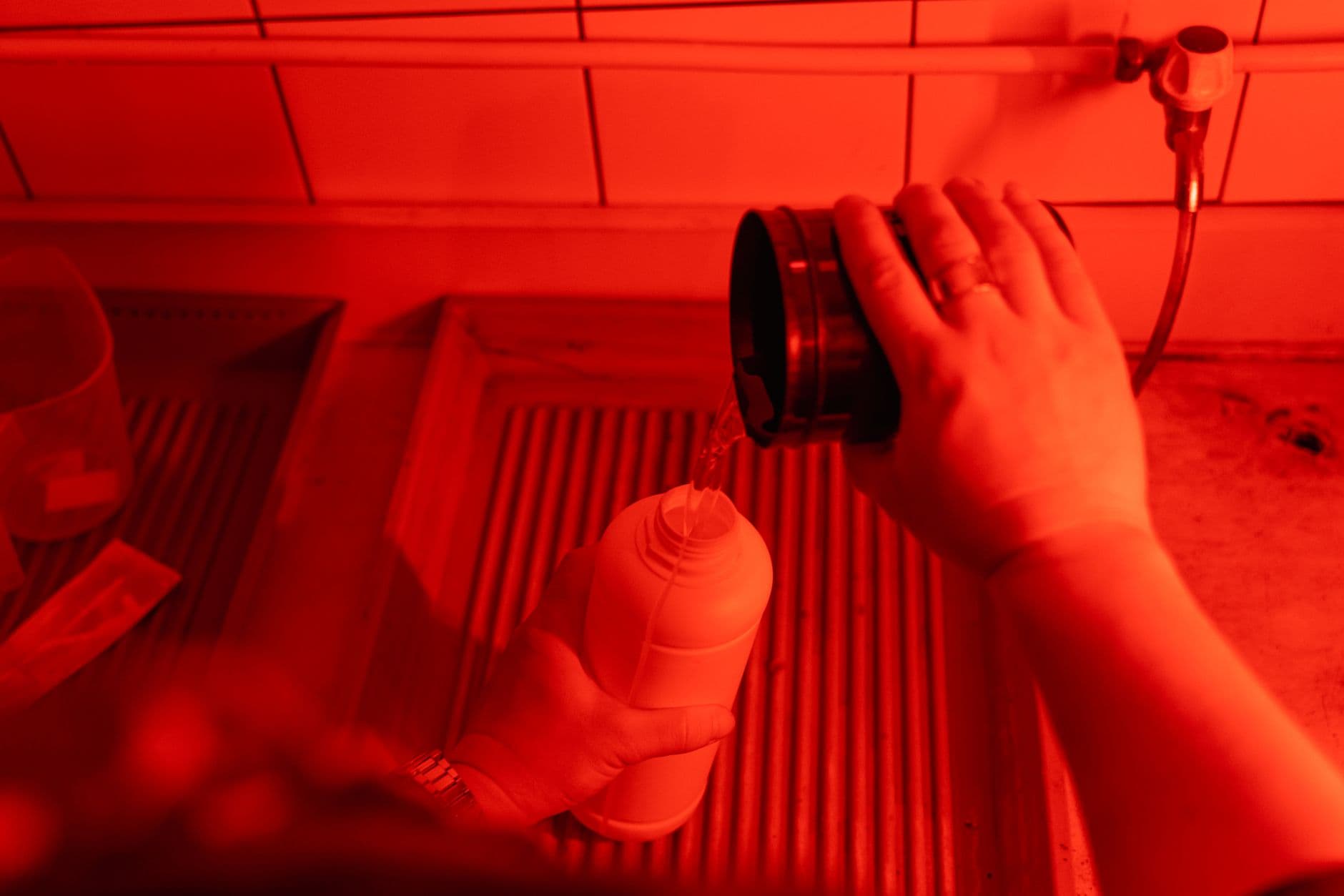 Person handling film-development chemicals under red safelight.
